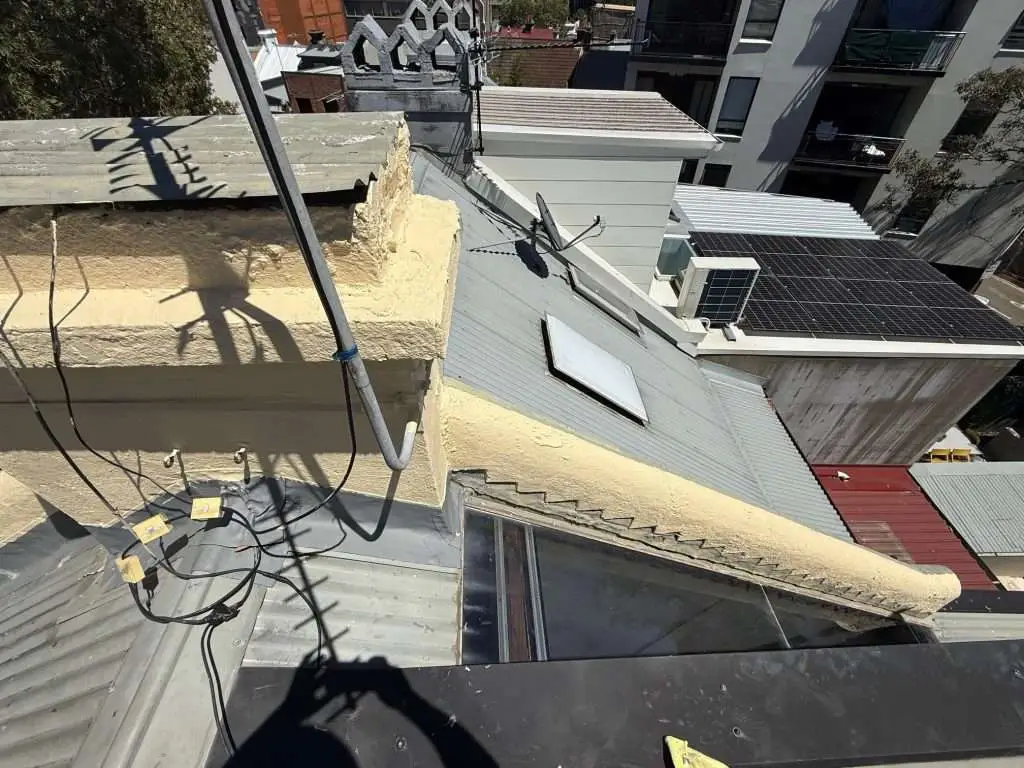 Aerial shot of damaged parapet wall on grey ribbed metal roof, crumbling plaster with downpipe