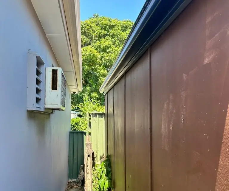 Side view of house gutter and fascia installation, grey metal edge with white downpipe, brown fence and trees in backyard
