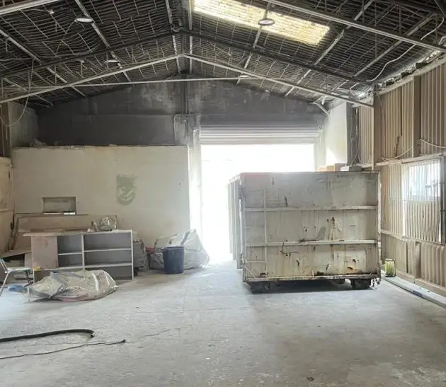 Inside view of industrial shed during construction or renovation, exposed metal roof structure, dumpster bin and concrete floor
